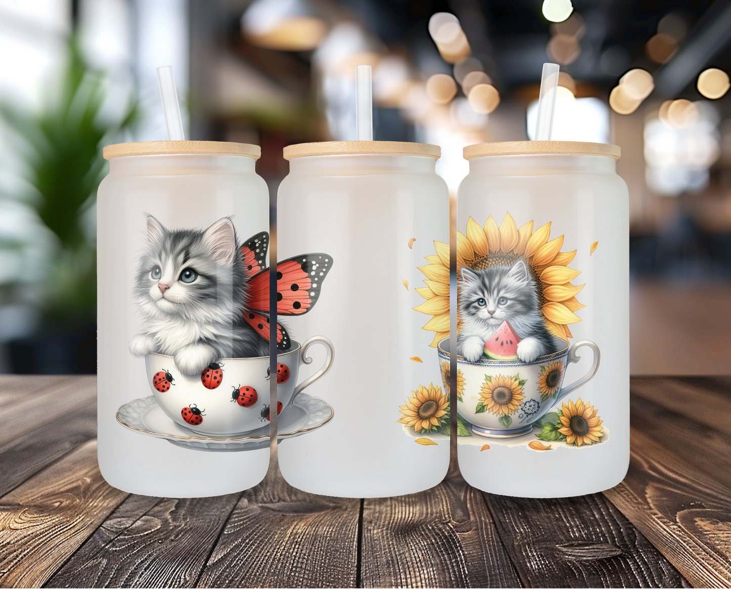 Three frosted glass cans with kitten designs, displayed on a wooden table against a blurred cafe background. One can features a kitten with ladybugs, and the other a kitten in a sunflower cup, both topped with bamboo lids and clear straws