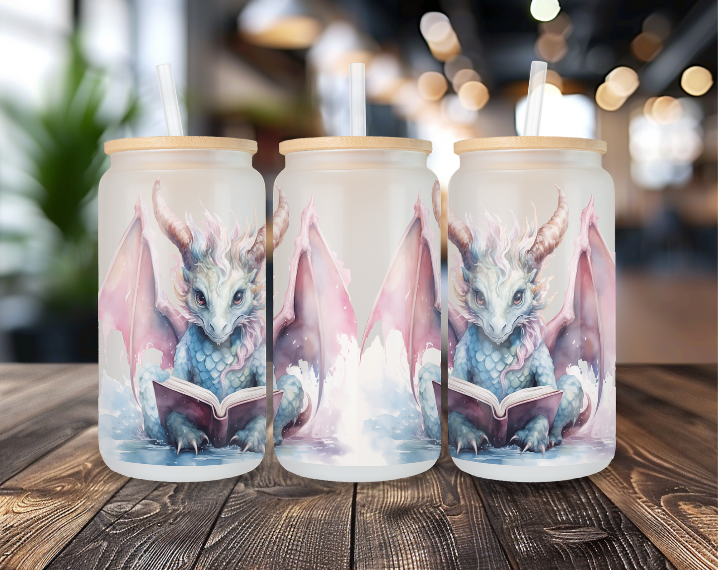 Three different perspectives of a frosted glass can featuring a mystical dragon with horns, pink and blue scales, and large wings, reading a book. Each glass has a bamboo lid and a clear straw, displayed on a wooden table with a blurred indoor background