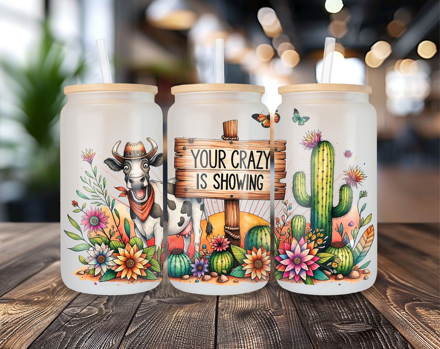 Three different perspectives of a frosted glass can featuring a Western-themed design: a cow in a cowboy hat, a rustic sign reading "Your Crazy is Showing," and a colorful cactus garden. Each glass has a bamboo lid and clear straw, displayed on a wooden table with a blurred indoor background.