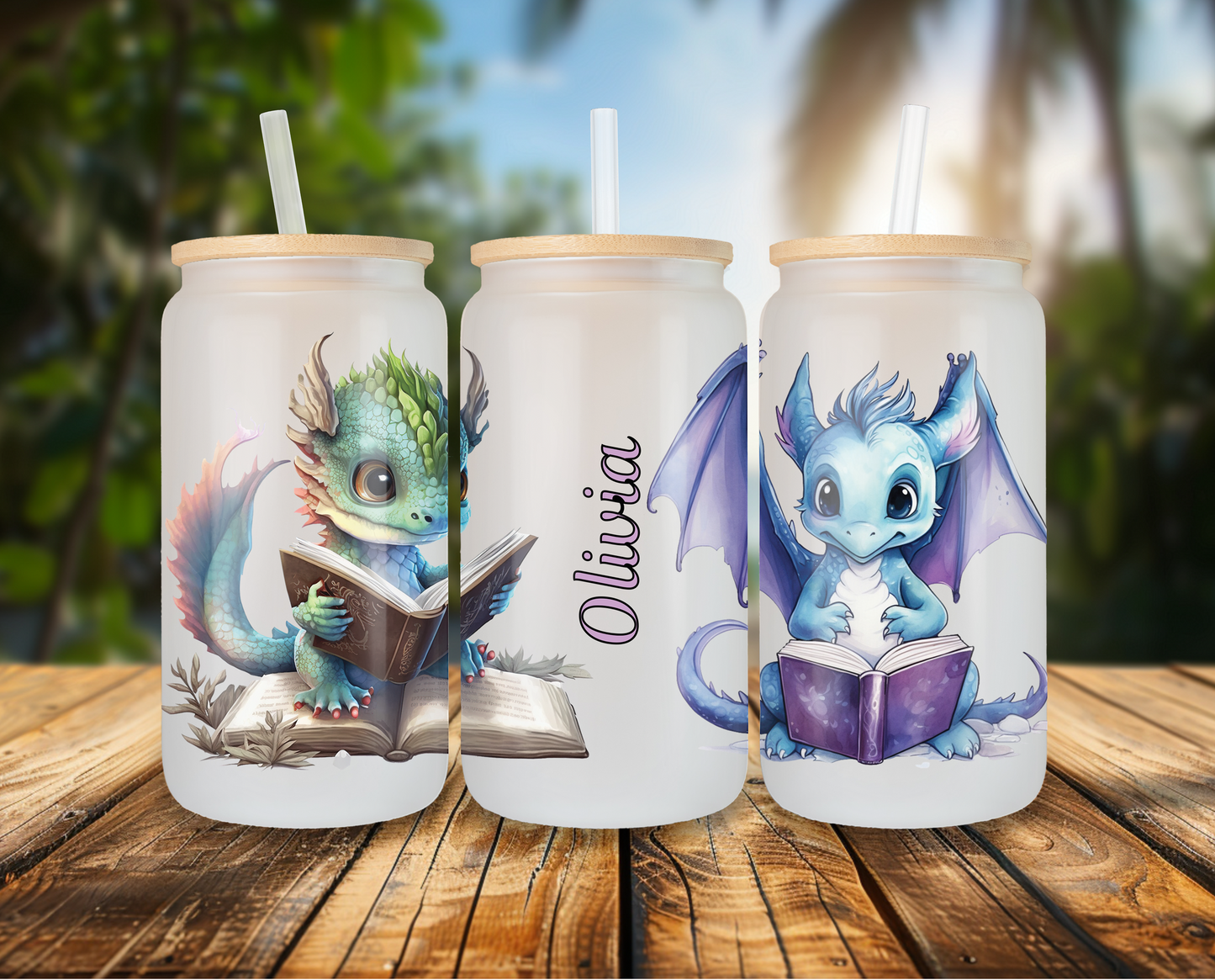 Three frosted glass cans with customizable dragon designs, one green and one blue, each reading a book, displayed on a wooden table with tropical background. Names 'Olivia' appear on one side of the can, topped with bamboo lids and clear straws.