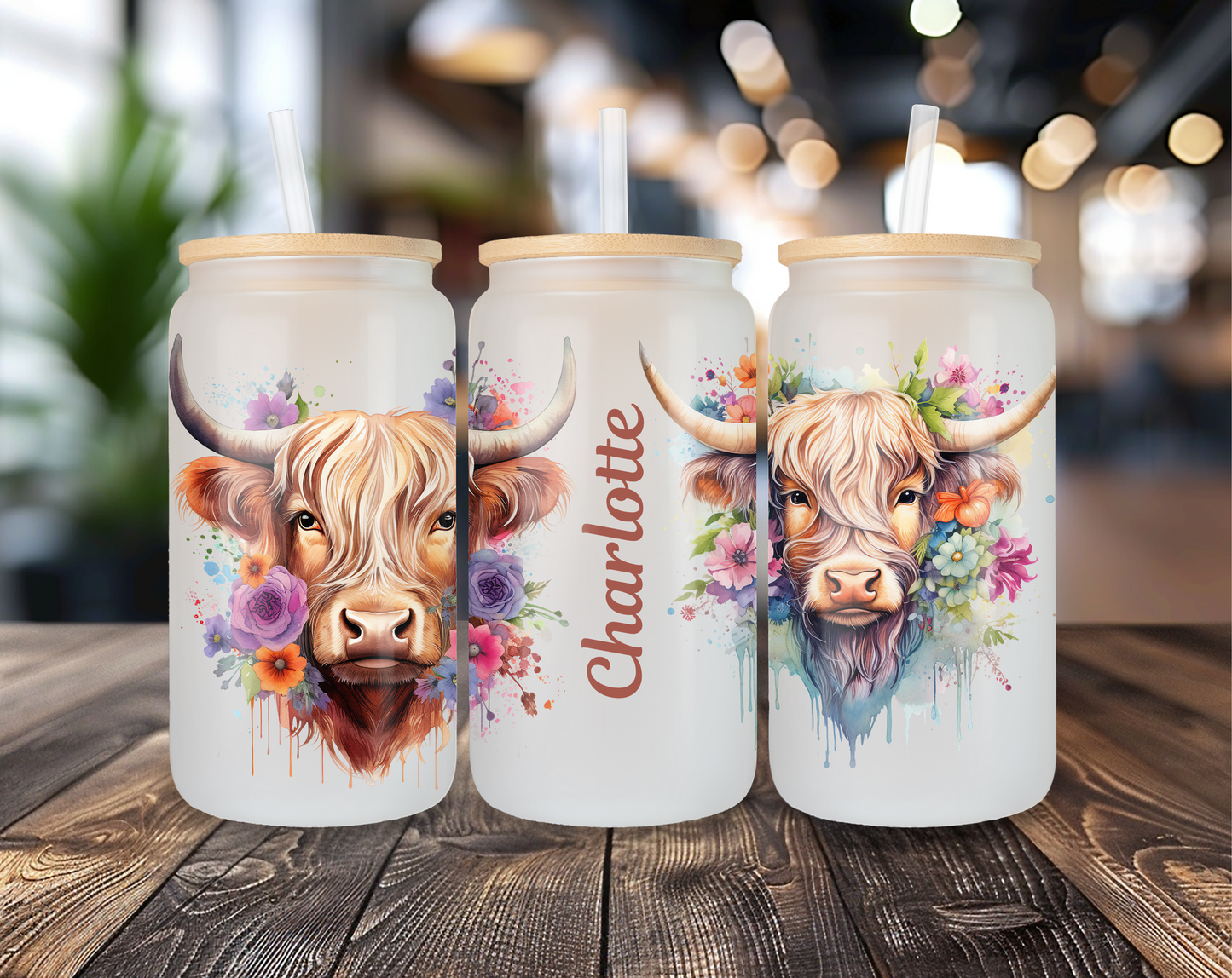 Three different perspectives of a frosted glass can featuring a Highland cow with floral accents. One perspective includes a customizable name, displayed as "Charlotte." Each glass has a bamboo lid and clear straw, set on a wooden table with a blurred indoor background.