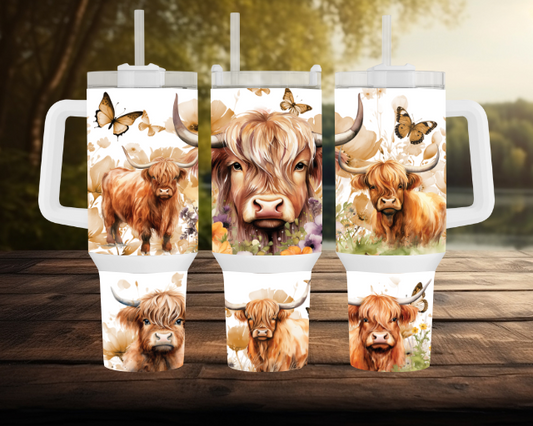 Three 40OZ tumblers with floral Highland cow designs, butterflies, and white lids with straws, displayed on a wooden table with a scenic lake and forest in the background.