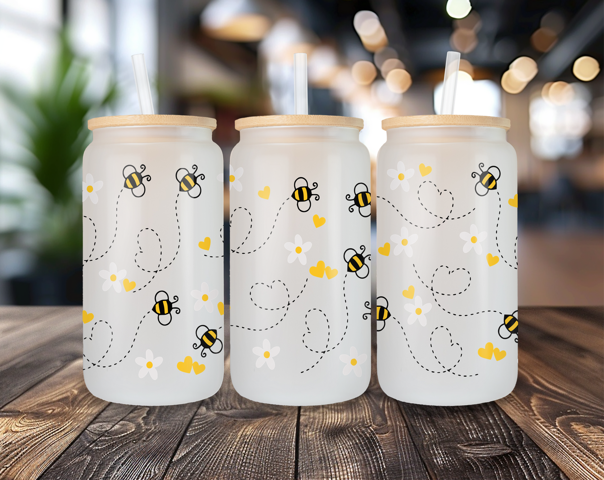 Three different perspectives of a frosted glass can featuring a playful bee design with daisies and yellow hearts. Each glass has a bamboo lid and a clear straw, displayed on a wooden table with a blurred indoor background.