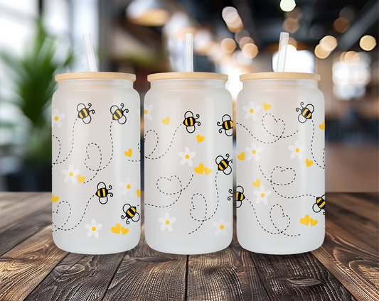 Three different perspectives of a frosted glass can featuring a playful bee design with daisies and yellow hearts. Each glass has a bamboo lid and a clear straw, displayed on a wooden table with a blurred indoor background.