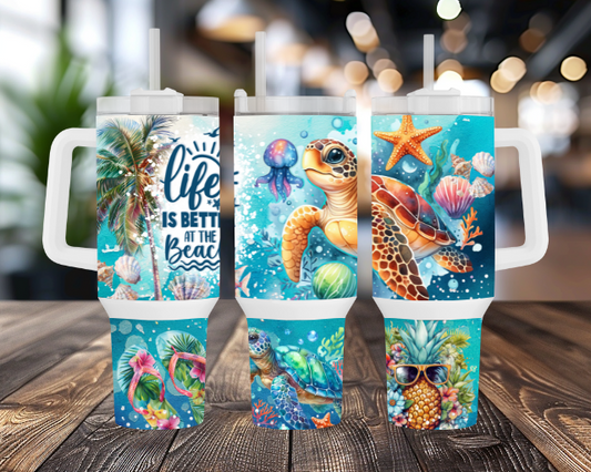 Three 40OZ tumblers featuring tropical sea turtles, jellyfish, seashells, palm trees, and a "Life is Better at the Beach" quote, with white lids and straws, on a wooden table and blurred indoor background.