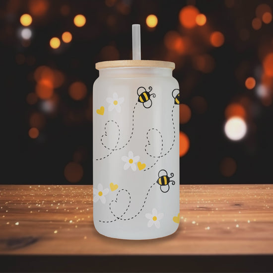 Video of Three different perspectives of a frosted glass can featuring a playful bee design with daisies and yellow hearts. Each glass has a bamboo lid and a clear straw, displayed on a wooden table with a blurred indoor background.