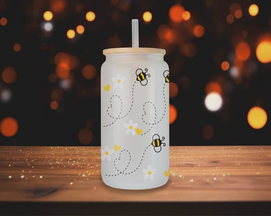 Video of Three different perspectives of a frosted glass can featuring a playful bee design with daisies and yellow hearts. Each glass has a bamboo lid and a clear straw, displayed on a wooden table with a blurred indoor background.